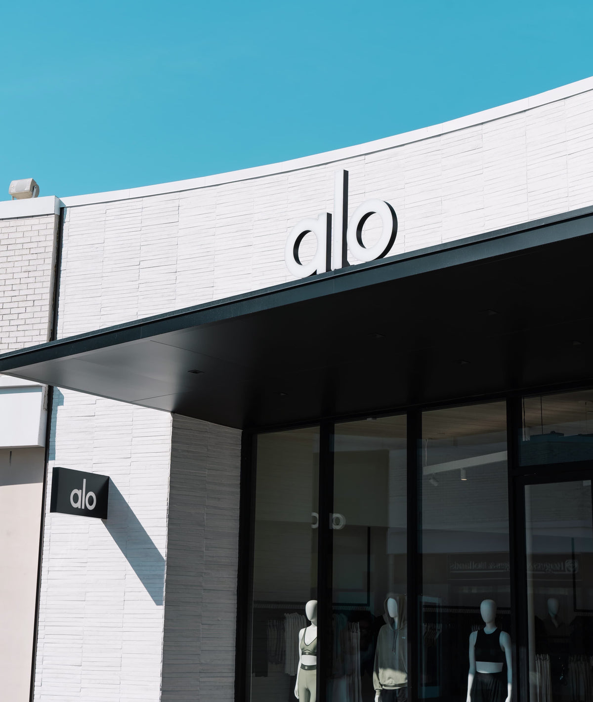 A photo of the front of the Alo Yoga Old Orchard store from a right angle showcasing the matte black awning and white stone façade with floor-to-ceiling front slider doors.