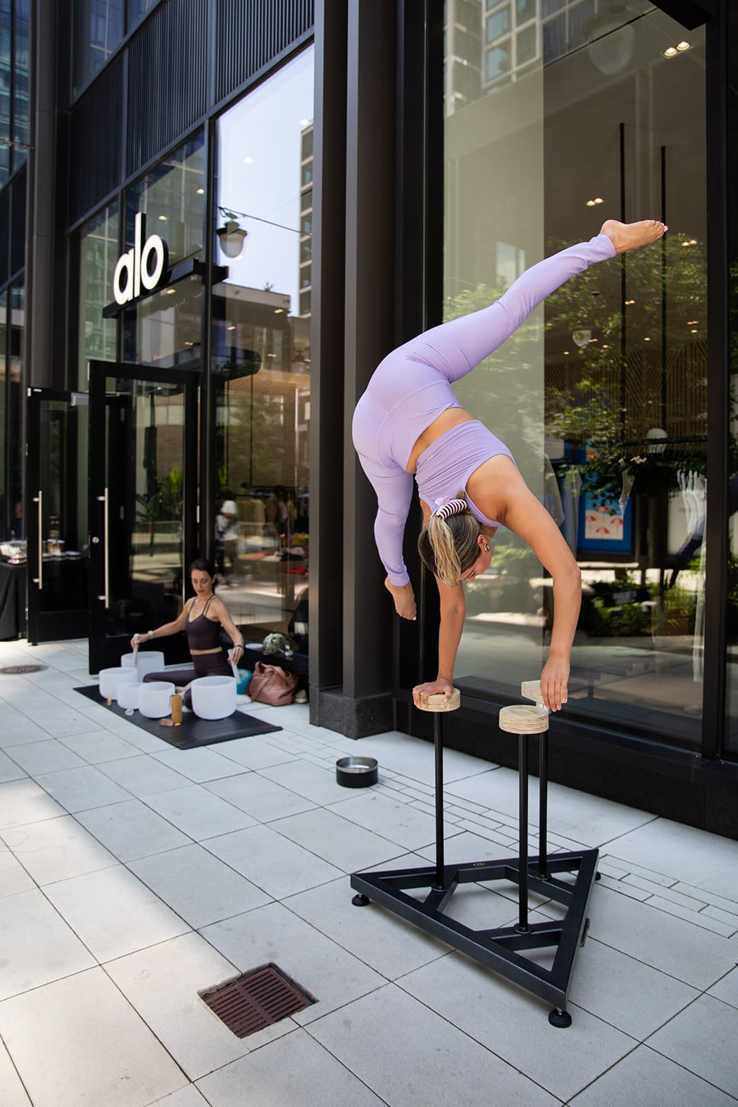 A woman doing acro yoga on two elevated blocks in front of the Alo Boston storefront with a woman in the background performing a sound bath with sound bowls.