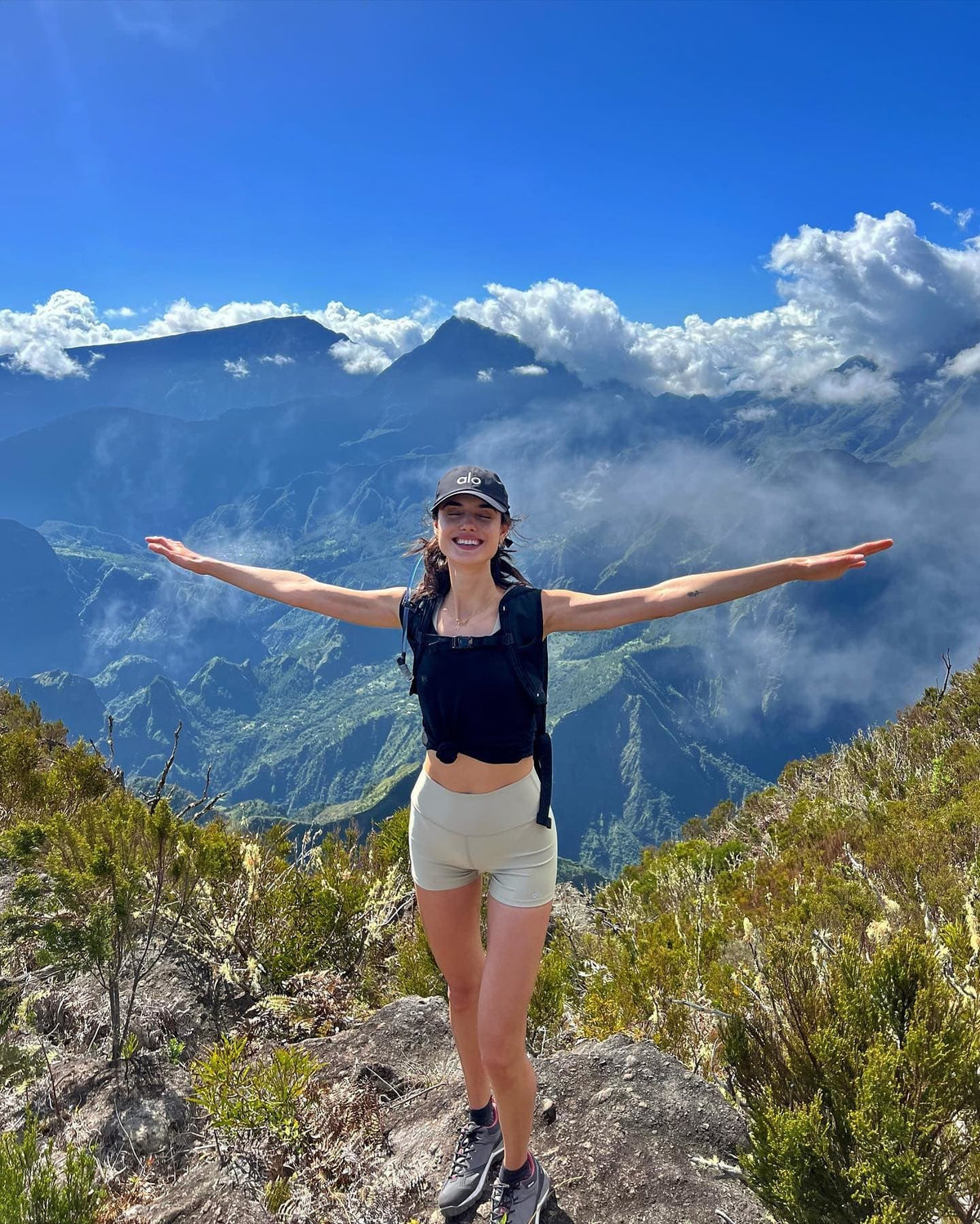 @blancapadilla wearing a pair of workout shorts and a black workout tank-top while posing high on a mountain during a hike with a large mountain range in the background and clouds peaking through the peaks in the distance.