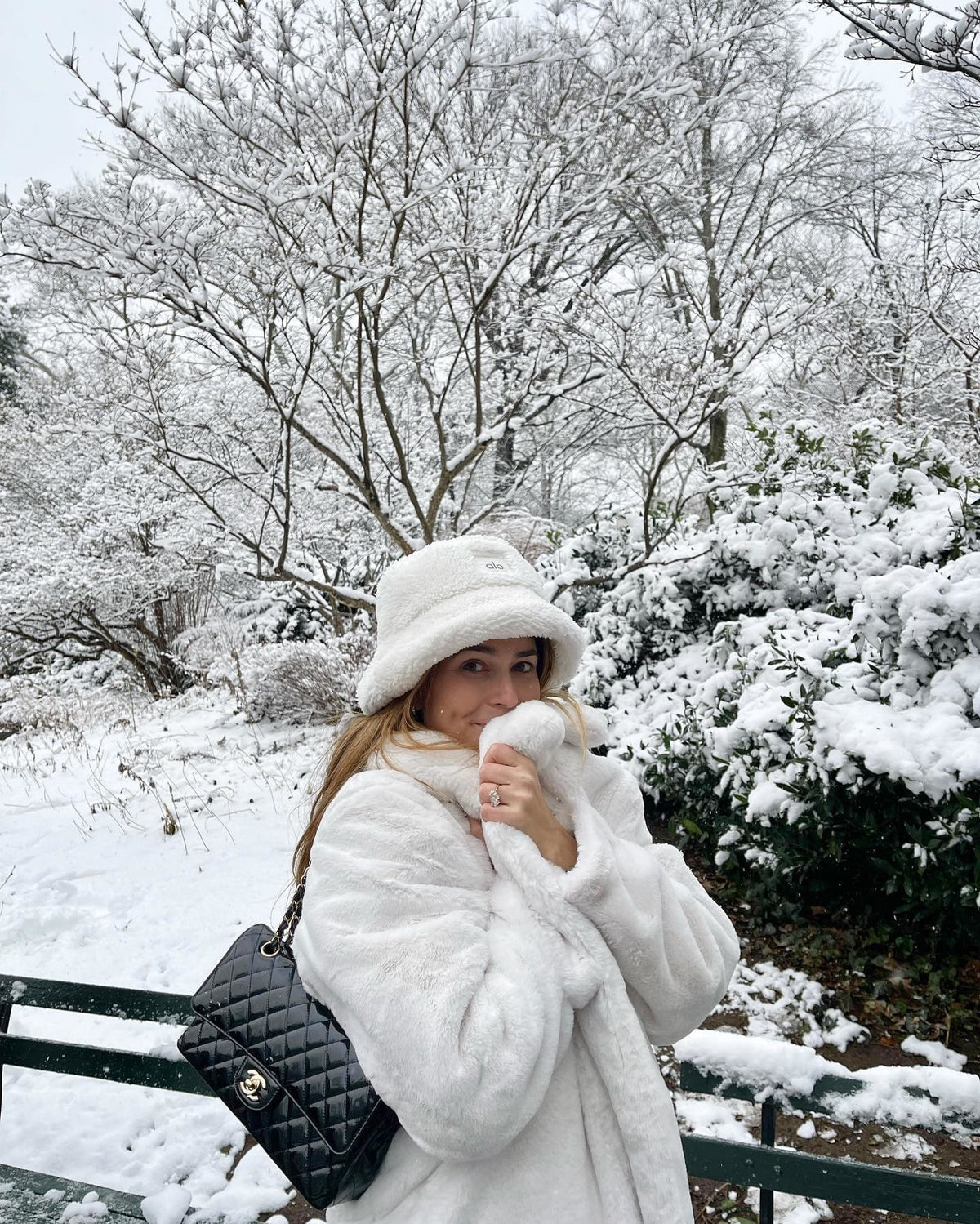 @coralsimanovich wearing a white faux fur trench coat with a sherpa bucket hat and a Chanel purse while walking a snowy park.