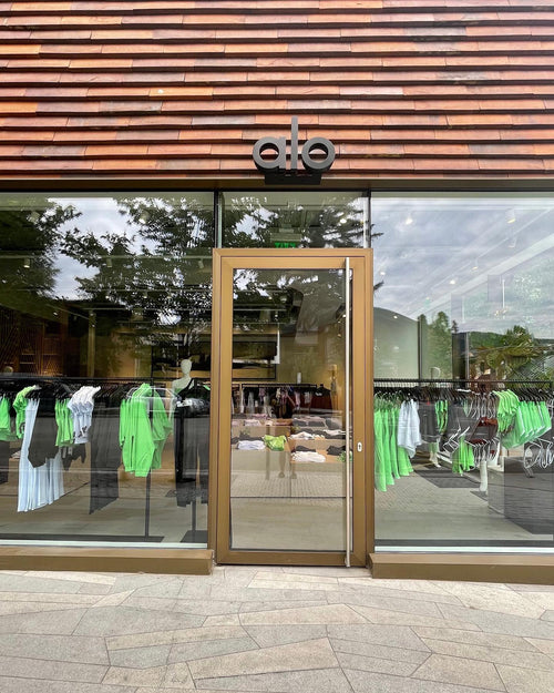 A photo of Alo Aspen’s storefront displaying wooden slats on the top, floor-to-ceiling windows beside the glass door, and an array of neon green apparel displayed in the windows.